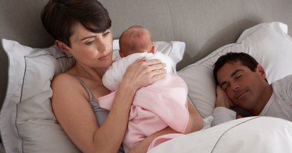 Mutter angestrengt mit Baby im Bett neben schlafendem Vater