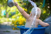 Kleinkind sitzt draußen in blauer Wanne und spritzt mit Wasser.