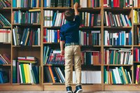 Junge im Grundschulalter stehr vor riesiger Bücherwand und sucht ein Buch aus.