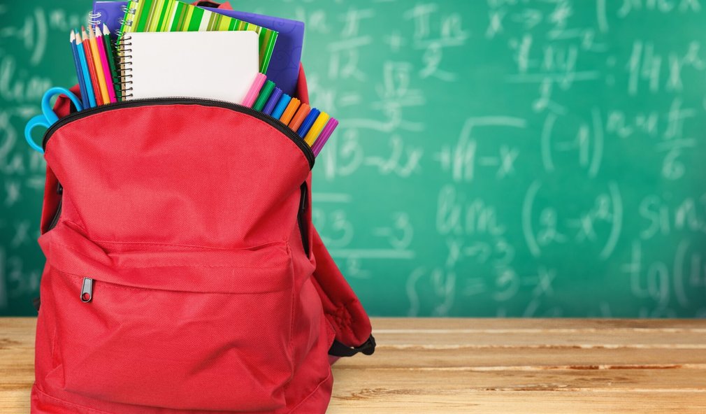 Roter Rucksack mit Schulsachen. Dahinter eine grüne Tafel mit Formeln drauf