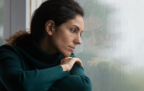 Traurige Frau sitzt mit Kopf am Fenster gelehnt und sieht durch Fensterscheibe mit Regntropfen.