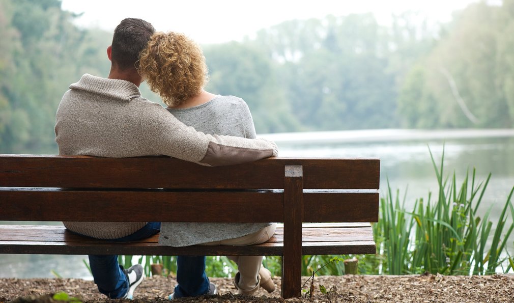 Frau und Mann sitzen liebevoll auf Bank. Schauen auf See.