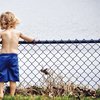 Kleiner Junge, nur Hose an, steht vor Zaun am See