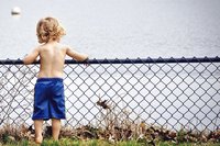 Kleiner Junge, nur Hose an, steht vor Zaun am See