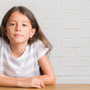 junges Mädchen sitzt am Tisch vor weißer Wand, schaut in Kamera.