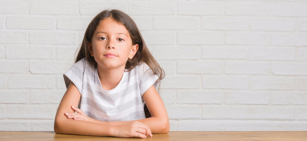junges Mädchen sitzt am Tisch vor weißer Wand, schaut in Kamera.