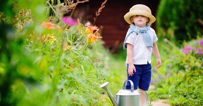 Kleinkind steht im Garten hat großen Hut auf mit Gießkanne