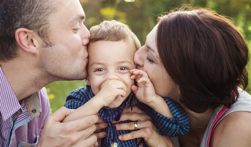 Kleines Kind wird von Eltern jeweils links und rechts auf die Wange geküsst.