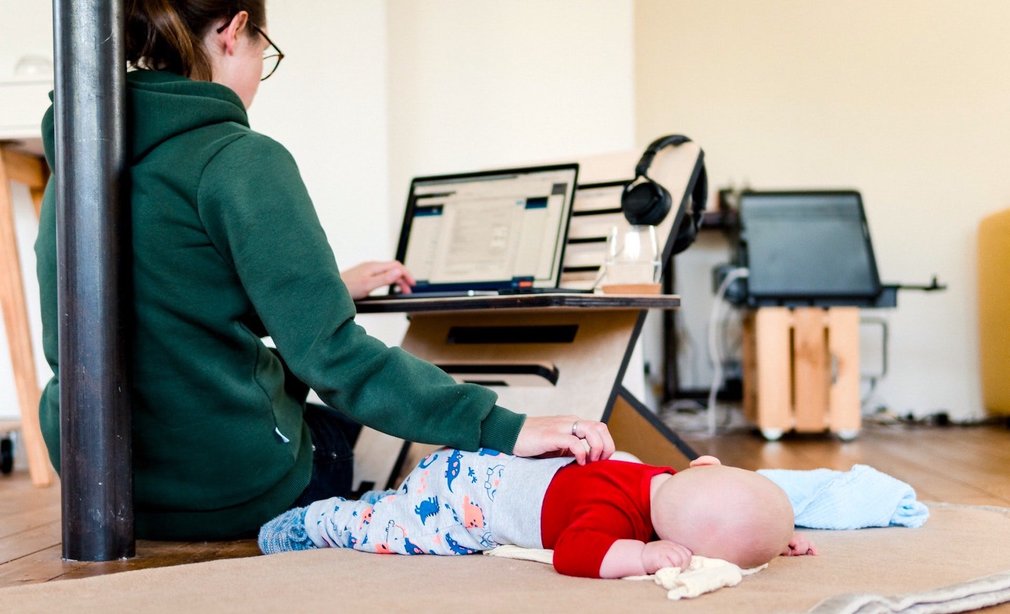 Mutter arbeitet am Laptop, Baby schläft auf Decke neben ihr. Sie hält Hand aufs Baby.