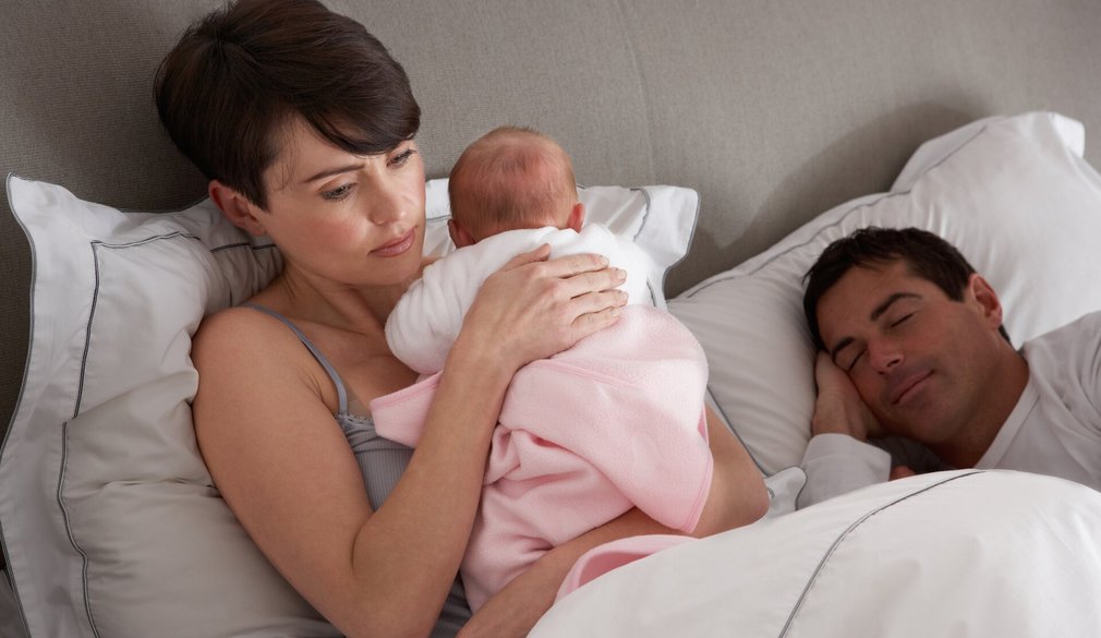 Mutter angestrengt mit Baby im Bett neben schlafendem Vater